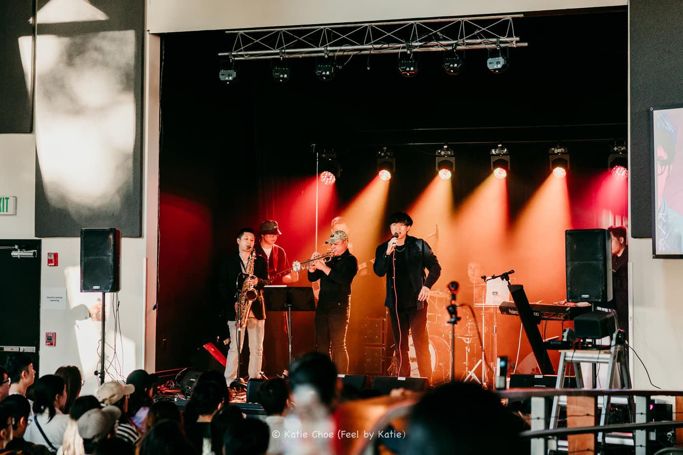 Band with saxophone, trumpet, and vocalist performing under red and amber beams from a truss rig, audience in the foreground