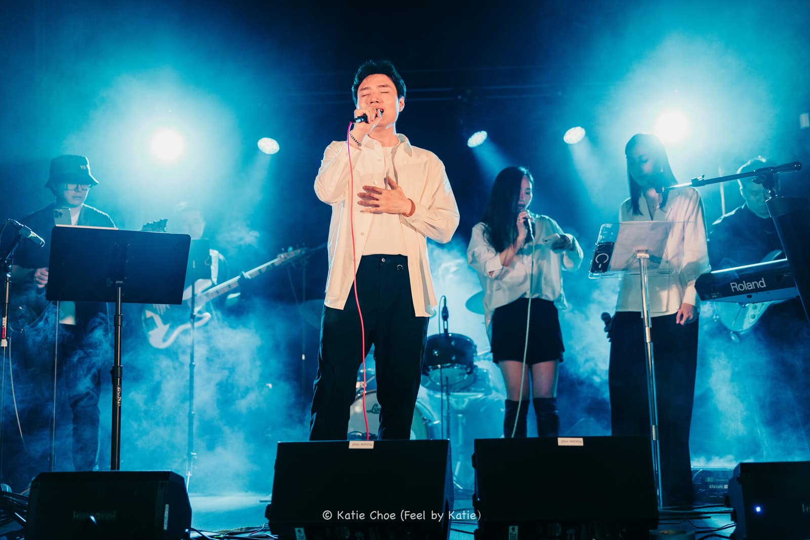 Vocalist singing centerstage under a striking blue and violet light wash, symmetrically flanked by the full band, at a Cuebeam-lit live show