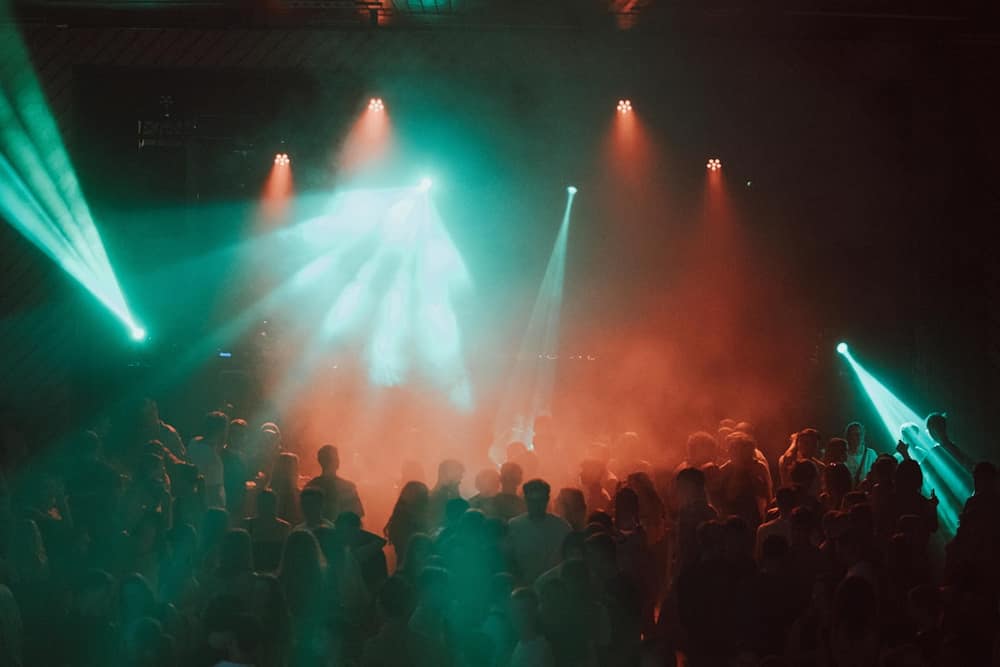 A crowd dancing under teal and red beams cutting through haze